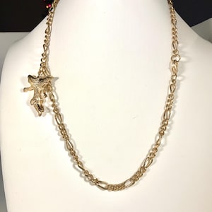 May include: A gold-colored necklace with a starfish pendant. The necklace features a chain with alternating oval and elongated links. The starfish pendant is detailed and hangs from a smaller chain.