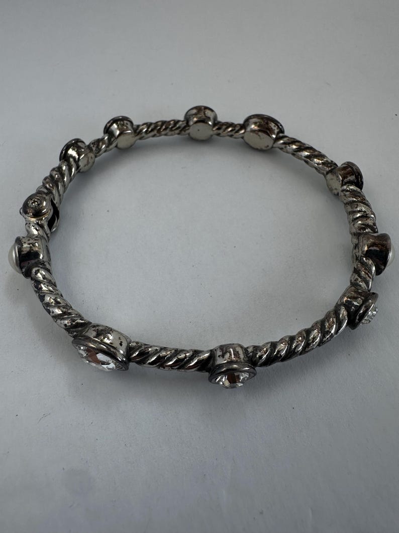 May include: Silver bangle bracelet with a twisted rope design. The bracelet has small, round, clear crystal accents. The bracelet is circular and appears to be made of metal.