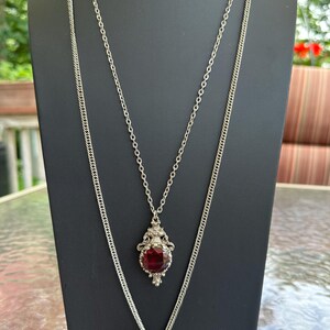 May include: A layered silver-tone necklace featuring two chains. The shorter chain holds an ornate silver-tone pendant with a deep red faceted oval stone. The longer chain features a round, coin-style pendant with a metallic finish and embossed detail.
