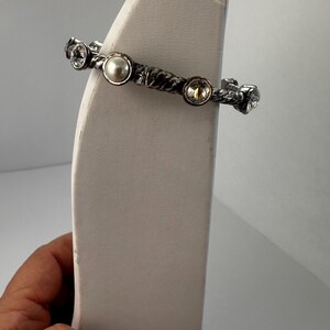 May include: Silver bracelet with a textured band and decorative embellishments. The bracelet features a central pearl and several clear and yellow crystals. The jewellery is displayed on a white stand.