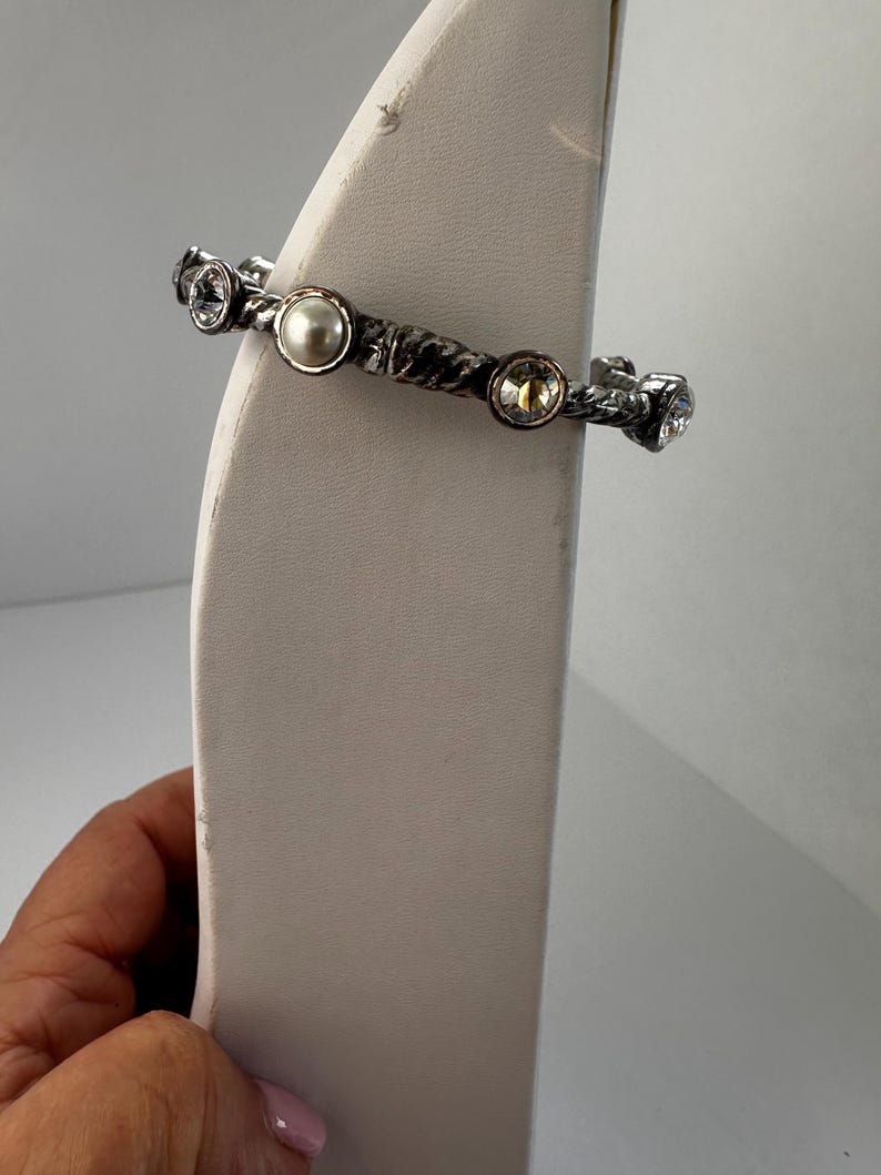 May include: Silver bracelet with a textured band and decorative embellishments. The bracelet features a central pearl and several clear and yellow crystals. The jewellery is displayed on a white stand.