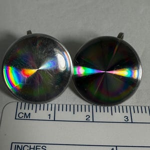 May include: Two silver-toned earrings with iridescent, rainbow-like finishes. The earrings are round and have a slightly domed shape. They measure approximately 1 inch in diameter.