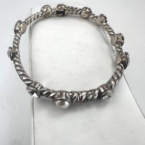 May include: Silver-coloured bangle bracelet with a twisted rope design. The bracelet features alternating embellishments of clear crystals, faux pearls, and small, round metal accents. The bracelet is a piece of jewellery.