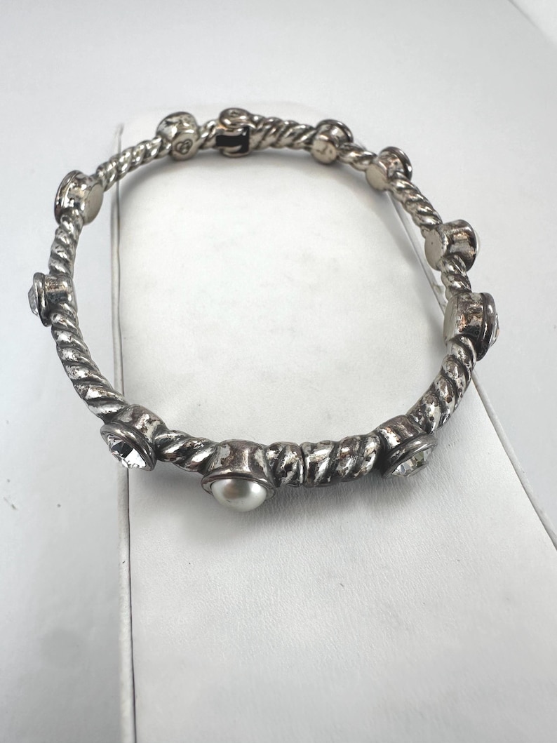 May include: Silver-coloured bangle bracelet with a twisted rope design. The bracelet features alternating embellishments of clear crystals, faux pearls, and small, round metal accents. The bracelet is a piece of jewellery.