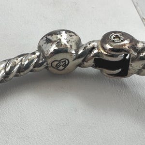 May include: Close-up of a silver bracelet clasp. The bracelet has a twisted rope design. The clasp features a decorative heart emblem and a secure locking mechanism. The metal shows signs of wear.