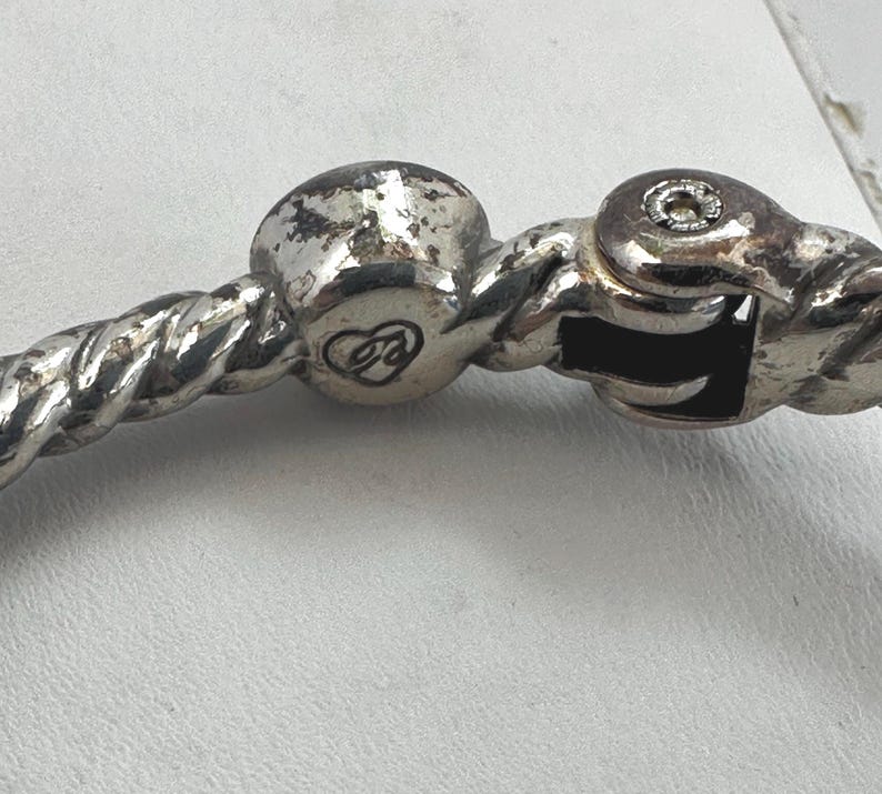 May include: Close-up of a silver bracelet clasp. The bracelet has a twisted rope design. The clasp features a decorative heart emblem and a secure locking mechanism. The metal shows signs of wear.