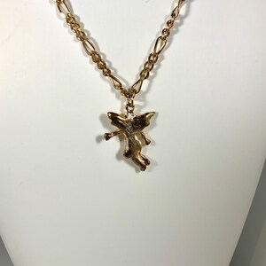 May include: A gold-colored necklace with a pendant shaped like a small angel. The necklace chain has a linked design. The angel pendant is detailed with wings and outstretched arms, giving it a dynamic appearance.
