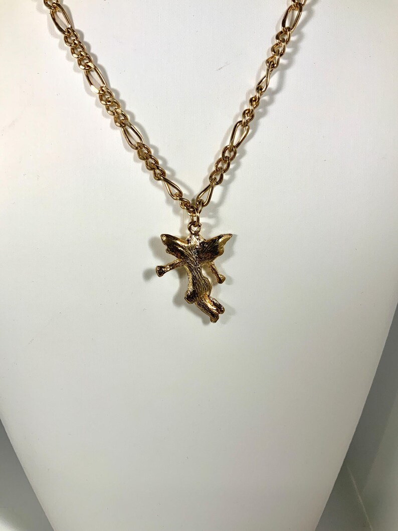 May include: A gold-colored necklace with a pendant shaped like a small angel. The necklace chain has a linked design. The angel pendant is detailed with wings and outstretched arms, giving it a dynamic appearance.