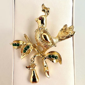 May include: A gold-tone brooch featuring a bird perched on a branch with green leaves and a pear hanging from the branch. The bird has red eyes and is decorated with gold details. The leaves are adorned with green rhinestones.