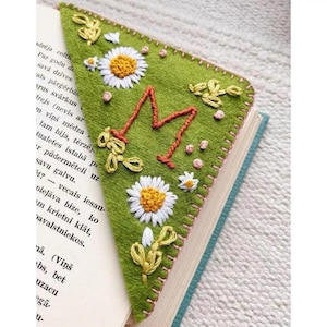 May include: A green felt bookmark with a large embroidered letter "M" in red thread. The bookmark is decorated with white daisies, pink dots, and green leaves.