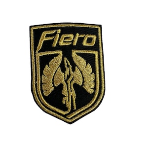 Puede incluir: Parche bordado con la palabra "Fiero" en letras doradas. El parche presenta una forma de escudo negro con un borde dorado y un diseño de fénix dorado en el centro. El parche es probablemente para ropa o accesorios.