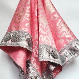 Może przedstawiać: Różowa tkanina ze złotym kwiatowym wzorem i srebrnym wykończeniem z cekinami. Tkanina jest udrapowana, ukazując skomplikowany wzór i dekoracyjne frędzle. Wykończenie ma srebrne cekiny i kwiatowe akcenty.