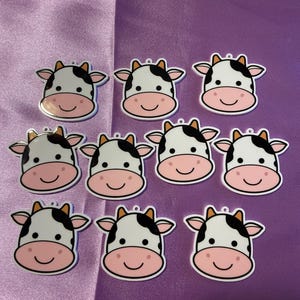 以下が含まれることがあります： 茶色の耳とピンクの鼻を持つ白黒の牛の頭の形をした樹脂製のチャームが10個。チャームの上部に吊り下げ用の穴があります。