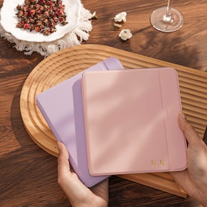 May include: Two pastel-colored tablet cases, one lavender and one pink, are held in a person's hands. The pink case has gold initials. A wooden tray, a white plate with dried flowers, and a wine glass are also visible.