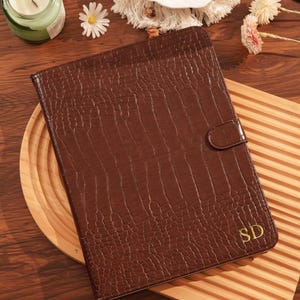 May include: A brown leather journal with a crocodile skin texture. The journal has a gold-colored clasp and the initials "SD" in gold. The journal is on a wooden tray with a candle and flowers in the background.