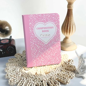 Puede incluir: Cuaderno de composición rosa con un corazón blanco y el texto "COMPOSITION BOOK Custom" en la portada. El cuaderno está sobre una estera de paja tejida.