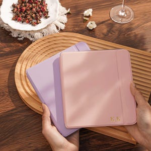 Op de afbeelding: Twee pastelkleurige tablethoezen, één in lavendel en één in roze, worden getoond. De roze hoes heeft de gouden initialen "K.K." op de voorkant. De hoezen liggen op een houten dienblad. Een wit bord met gedroogde bloemen en een wijnglas zijn ook zichtbaar.