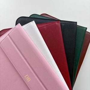 May include: A collection of colorful, leather-like tablet cases in various colors, including pink, white, red, black, green, and brown. The pink case has gold-colored initials "DB" on the front. The cases are arranged in a fan shape.