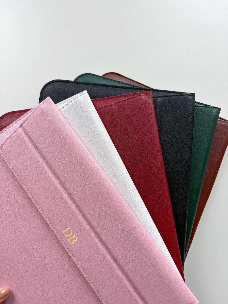 May include: A collection of colorful, leather-like tablet cases in various colors, including pink, white, red, black, green, and brown. The pink case has gold-colored initials "DB" on the front. The cases are arranged in a fan shape.