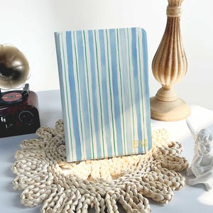 Może przedstawiać: Notatnik z niebiesko-zieloną okładką w paski stoi na plecionym podkładce. Notatnik ma złote inicjały. Świecznik w stylu vintage i mała statua znajdują się w tle.