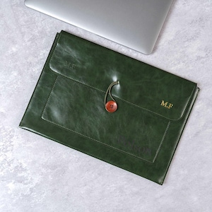 Puede incluir: Una funda para portátil de cuero verde oscuro con cierre de botón y un pequeño bolsillo. Las iniciales "M.F" están grabadas en la funda. La funda está sobre una superficie gris claro junto a un portátil.
