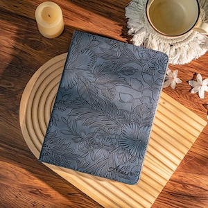 May include: A blue leather journal with a floral embossed design. The journal features a detailed pattern of leaves and flowers. The word "Hana" is written in cursive on the bottom right corner. The journal is on a wooden surface.