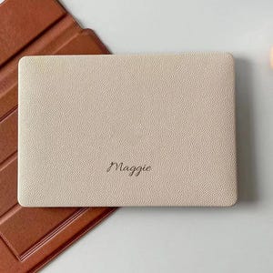 Puede incluir: Una funda rectangular para tableta de color crema con una superficie texturizada y granulada que presenta el nombre Maggie grabado en una escritura cursiva oscura. La funda está colocada parcialmente sobre una cubierta protectora marrón lisa.