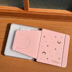 May include: A light pink tablet case featuring a gold foil celestial pattern of moons, stars, and plus signs. The cover includes the text Yikoco and a fold-out stand mechanism. A clear plastic frame with a protective film label is visible underneath.