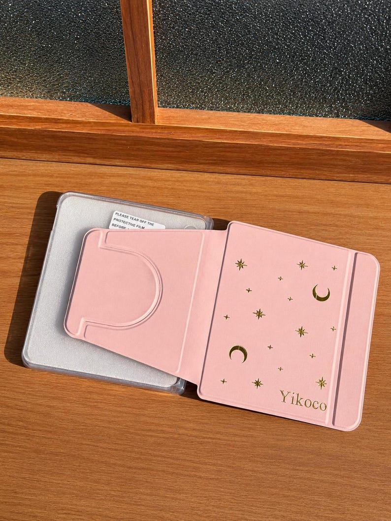 May include: A light pink tablet case featuring a gold foil celestial pattern of moons, stars, and plus signs. The cover includes the text Yikoco and a fold-out stand mechanism. A clear plastic frame with a protective film label is visible underneath.