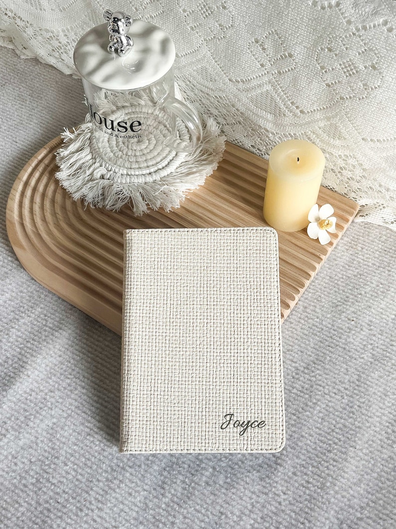 May include: A white, textured notebook with the name "Joyce" embossed in black lettering. The notebook is sitting on a wooden surface with a white candle and a white mug with a bear on top.