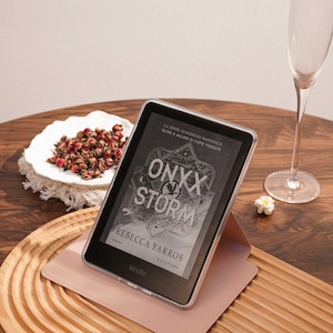 May include: A black Kindle e-reader in a clear protective case, resting on a light pink folding stand. The screen displays the book cover for Onyx Storm by Rebecca Yarros, featuring geometric line art and bold white text against a dark background.