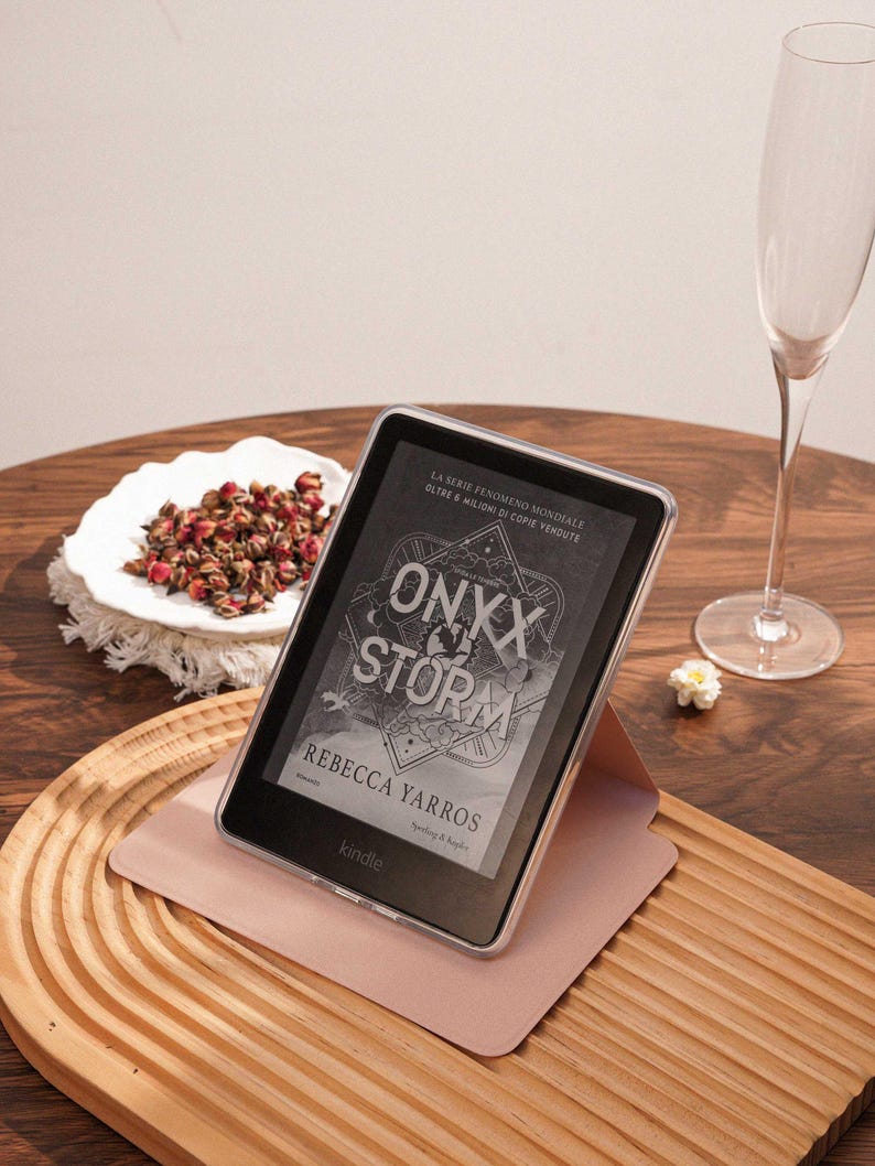May include: A black Kindle e-reader in a clear protective case, resting on a light pink folding stand. The screen displays the book cover for Onyx Storm by Rebecca Yarros, featuring geometric line art and bold white text against a dark background.