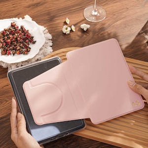 May include: A light pink tablet case with a foldable stand feature and the gold embossed initials AS in the bottom right corner. The case is shown partially open, revealing a dark gray tablet screen inside the protective frame.