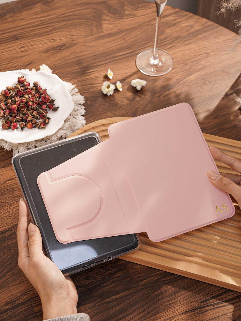 May include: A light pink tablet case with a foldable stand feature and the gold embossed initials AS in the bottom right corner. The case is shown partially open, revealing a dark gray tablet screen inside the protective frame.