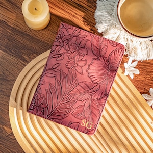 May include: A burgundy leather passport holder with a floral embossed design. The cover features detailed leaves and flowers, with the initials "SG" in the lower right corner. The passport holder is rectangular and appears to be closed.