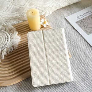 May include: A white tablet case with a textured woven design. The case is closed and lying on a wooden surface.