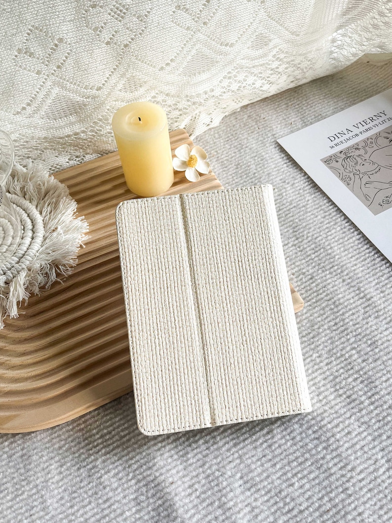 May include: A white tablet case with a textured woven design. The case is closed and lying on a wooden surface.