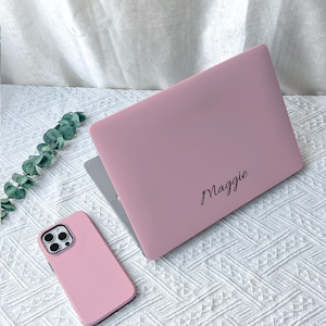 Puede incluir: Una funda rígida para portátil de color rosa mate con el nombre Maggie en letra cursiva negra, junto con una funda para smartphone de color rosa mate a juego. Ambos artículos tienen un acabado liso sobre una superficie blanca texturizada.