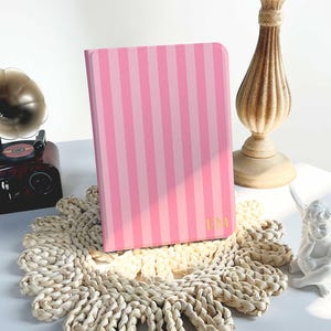 May include: A pink notebook with vertical stripes and gold initials. The notebook is propped up on a woven placemat, with a vintage gramophone and candle holder in the background. The notebook is a light pink color with darker pink stripes.