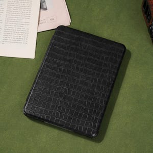 May include: Black rectangular case with a textured, crocodile-like pattern. The case is on a green surface, with a newspaper and a small electronic device in the background.