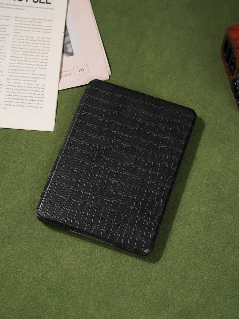 May include: Black rectangular case with a textured, crocodile-like pattern. The case is on a green surface, with a newspaper and a small electronic device in the background.