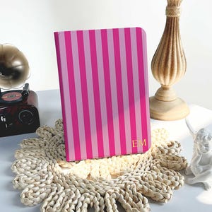 Puede incluir: Un cuaderno a rayas rosas y rosa claro con las iniciales doradas "EM" en la portada. El cuaderno descansa sobre un salvamanteles tejido. Un candelabro de estilo vintage y un pequeño gramófono están en el fondo.