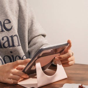 May include: A light pink tablet stand with a circular cutout design, supporting a tablet device at an angled position. The stand features a smooth, matte finish and a triangular base structure that provides stability for the electronic device.