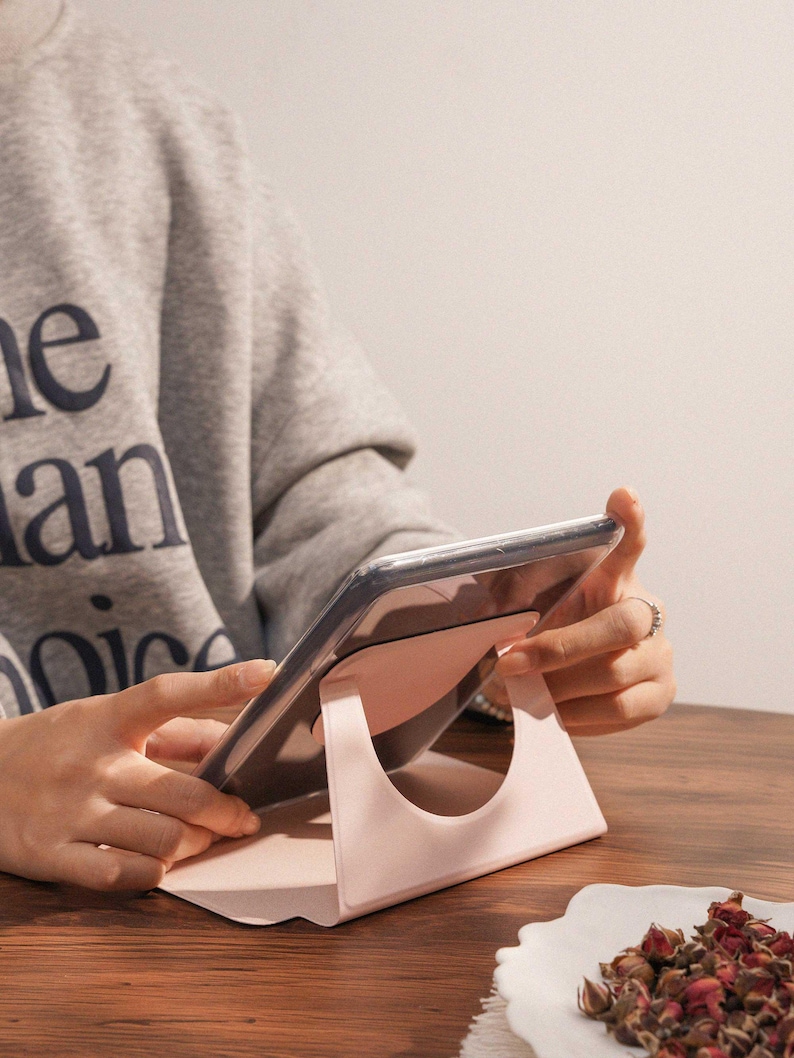 May include: A light pink tablet stand with a circular cutout design, supporting a tablet device at an angled position. The stand features a smooth, matte finish and a triangular base structure that provides stability for the electronic device.