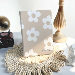 Pode incluir: Uma capa bege para tablet com um padrão de margaridas brancas e a palavra "Custom" em escrita preta. A capa está sobre um tapete de mesa tecido, com um castiçal decorativo e uma pequena estátua no fundo.