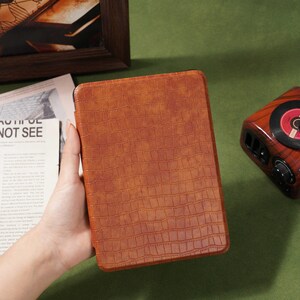 May include: A brown, textured tablet case with a crocodile skin pattern. The case is held in a person's hand. The background includes a framed picture, a vintage-style radio, and a newspaper with the text "NOT SEE".