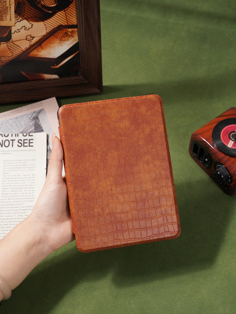 May include: A brown, textured tablet case with a crocodile skin pattern. The case is held in a person's hand. The background includes a framed picture, a vintage-style radio, and a newspaper with the text "NOT SEE".