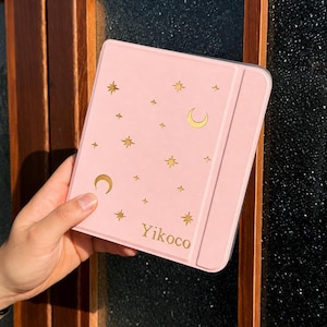 May include: A light pink tablet case featuring a gold foil pattern of crescent moons and various star shapes scattered across the front cover. The word Yikoco is printed in gold lettering near the bottom right corner of the rectangular protective cover.
