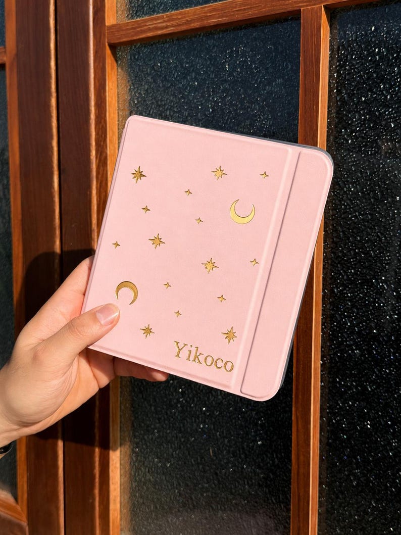 May include: A light pink tablet case featuring a gold foil pattern of crescent moons and various star shapes scattered across the front cover. The word Yikoco is printed in gold lettering near the bottom right corner of the rectangular protective cover.