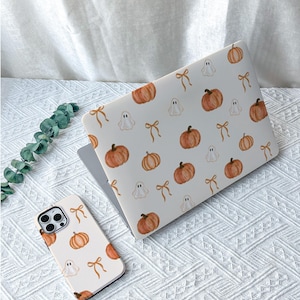 May include: A laptop and a smartphone case featuring a repeating pattern of orange pumpkins, white ghosts, and light brown bows on a white background. The design is consistent across both electronic device covers, showcasing a festive seasonal theme.